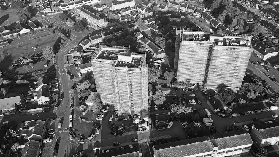 An aerial black and white photograph shows a multi-story residential building with a flat roof fitted with solar panels and various mechanical equipment. In front of the building, several wooden and c